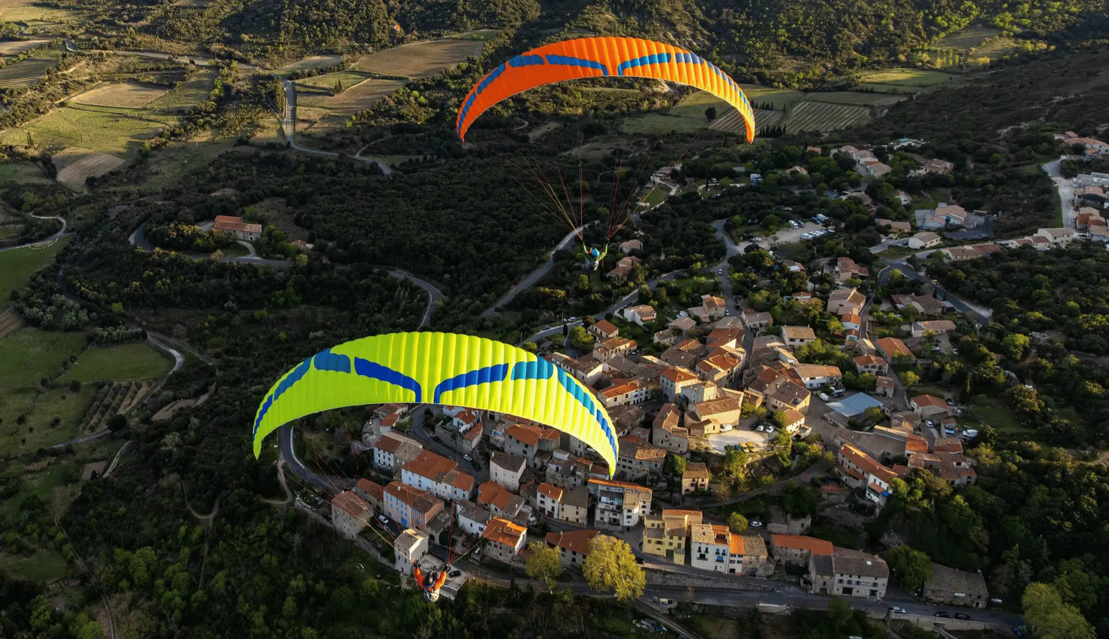 EN-B Gleitschirm-Klassen Vergleich: Zwei Paraglider, einer gelb-blau, der andere orange, gleiten über ein malerisches Dorf und grüne Hügel.