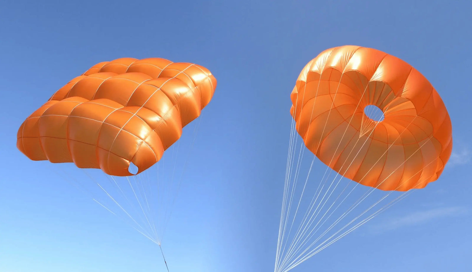 Vergleich von Gleitschirm-Rettungsschirmen: links eine Kreuzkappen-Rettung mit hoher Pendelstabilität, rechts eine klassische Rundkappen-Rettung mit zentraler Öffnung vor blauem Himmel
