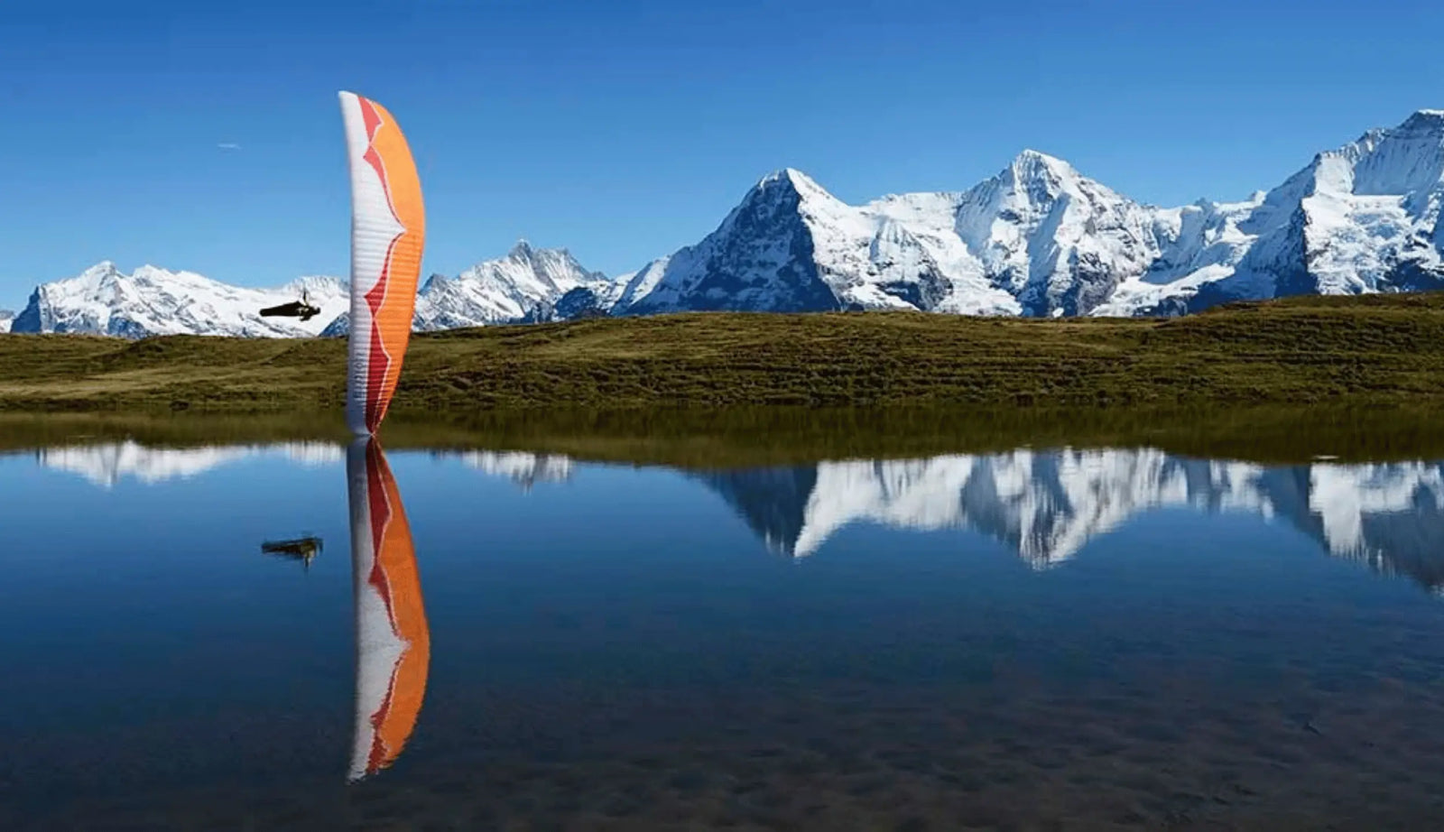 Orange und weiße Gleitschirme für 3-Leiner und 2-Leiner im Flug über einem Bergsee mit schneebedeckten Gipfeln.