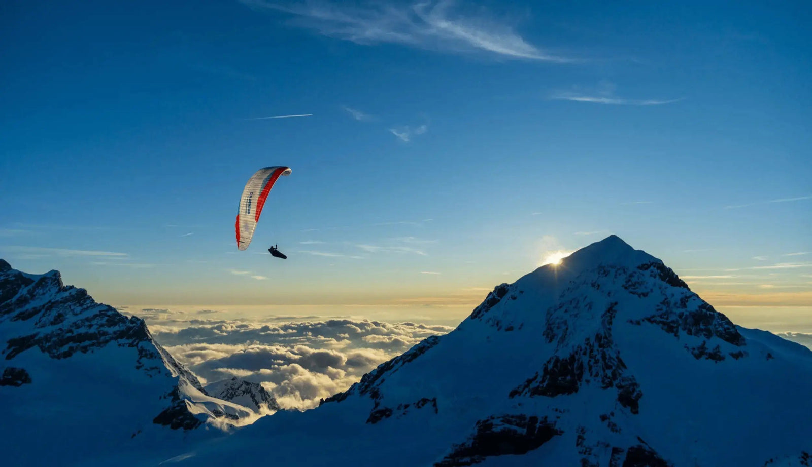 Ein Gleitschirmflieger schwebt über schneebedecktem Gebirgskamm bei Sonnenaufgang, unter ihm ein Wolkenmeer im Tal.