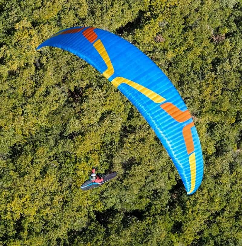 Ozone Swift 6 Gleitschirm beim Flug über Bäume, ideal für erfahrene Intermediate-Piloten. Leichtes, agiles Design mit hoher XC-Leistung und EN-B Zertifizierung.