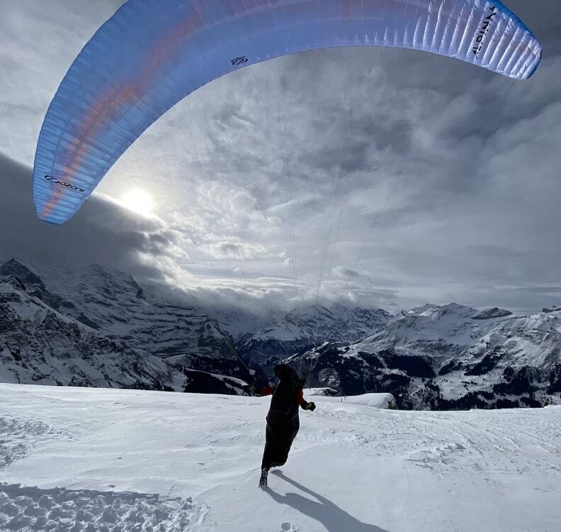 Person paragleitet mit Ozone Alpina 4 (EN-C) über schneebedecktem Berg, ideal für erfahrene Piloten, bietet hohe Leistung und leichtes Handling.