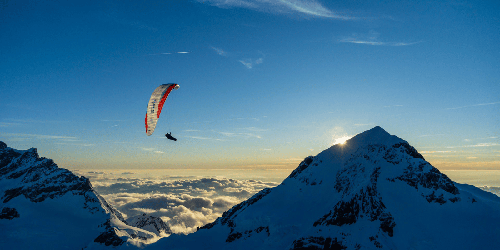 Paraglider fliegt bei Sonnenuntergang über schneebedeckte Berggipfel mit Wolkenmeer im Tal, Szene vermittelt Freiheit und Naturerlebnis beim Paragliding.