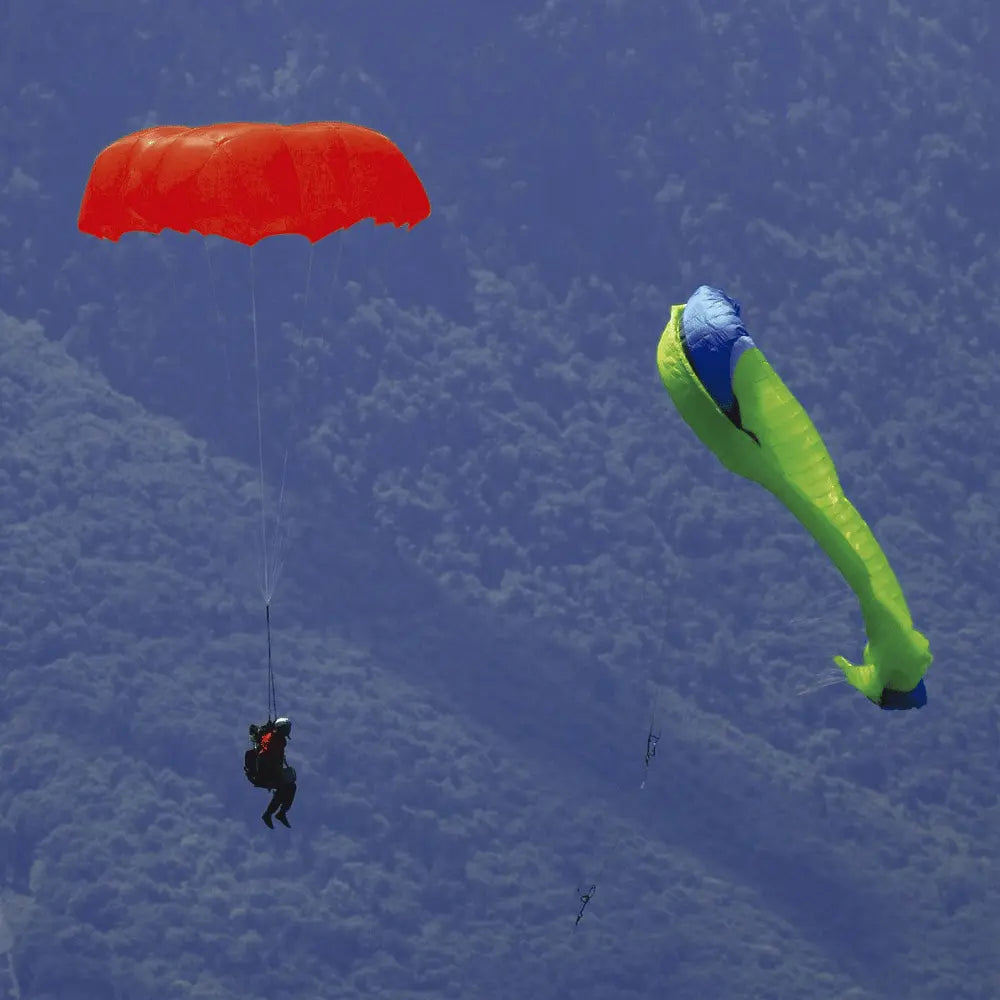 Person unter einem roten Niviuk Octagon 2 Rettungsschirm, sicher über bewaldeten Bergen schwebend, mit einem zweiten, teils eingeklappten Schirm daneben.