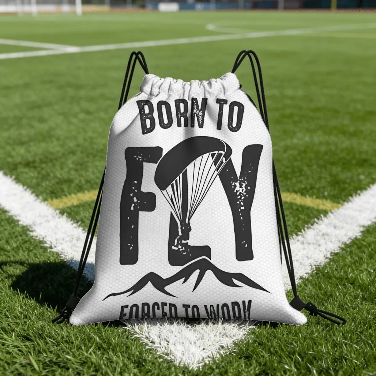 Weißer Sportbeutel mit Kordelzug, bedruckt mit dem Spruch "Born to Fly" und einer Paragliding-Silhouette, liegt auf einem Fußballfeld.