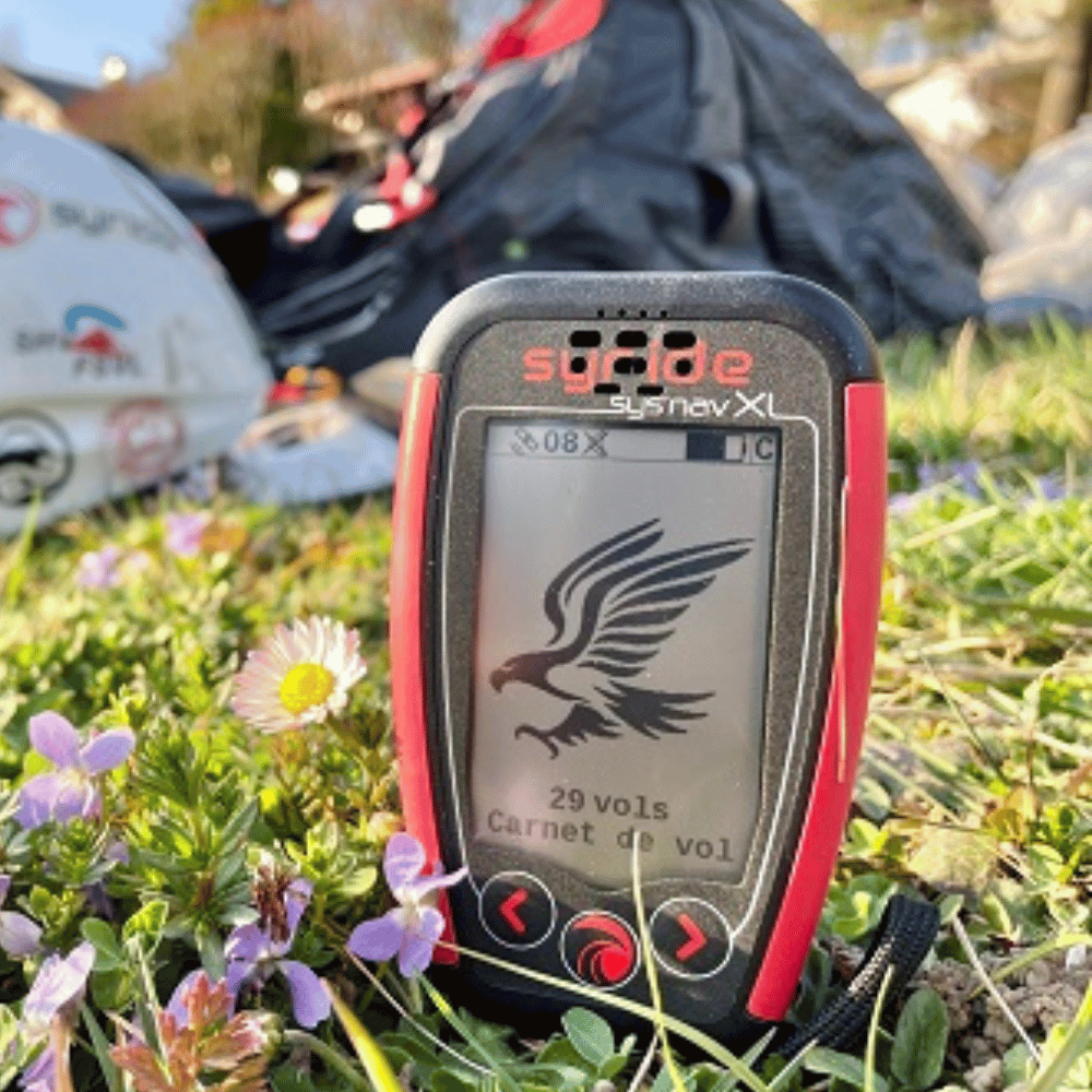 Syride SYS&#39;Nav XL - Kompaktes Fluginstrument im Gras und Gleitschirmausrüstung im Hintergrund, ideal für Paragliding-Enthusiasten, mit GPS, Bluetooth und weltweiter Kartierung.