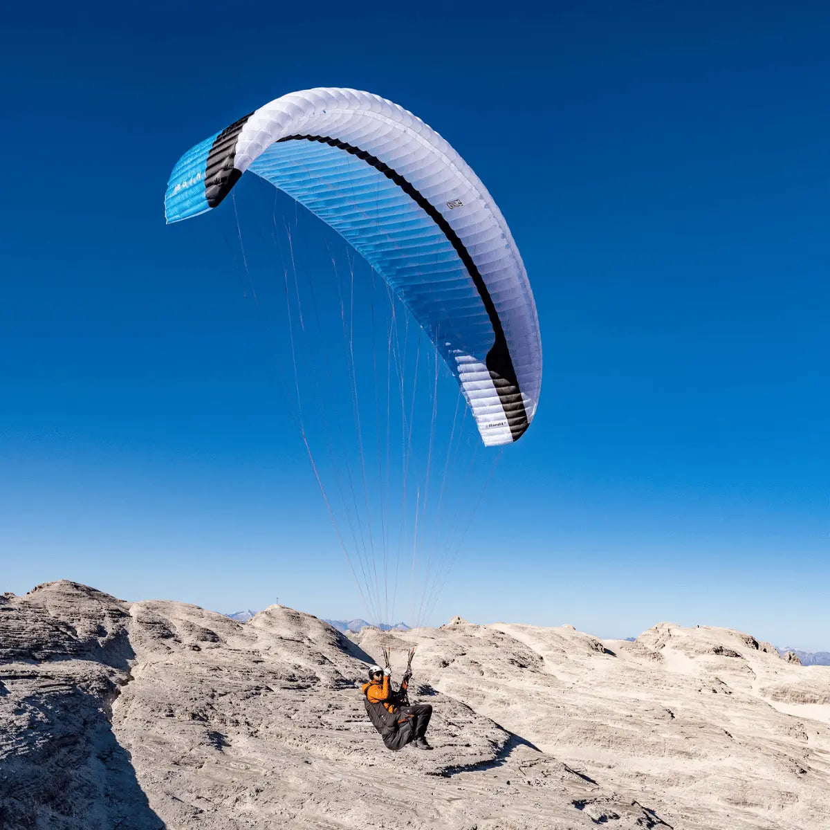 Gin Bandit 2 Ultra Light Gleitschirm über felsigem Grat, Pilot in orangefarbener Jacke, bereit zum Start. Perfekt für Thermik, XC und Hike & Fly.