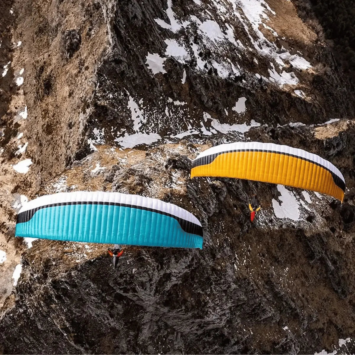Zwei Gleitschirme, ein türkisfarbener und ein gelber, gleiten elegant über eine felsige Berglandschaft mit Schneefeldern. Der türkisfarbene Gleitschirm hat eine weiße Oberseite und blaue Flügel, der gelbe eine weiße Oberseite und gelbe Flügel.