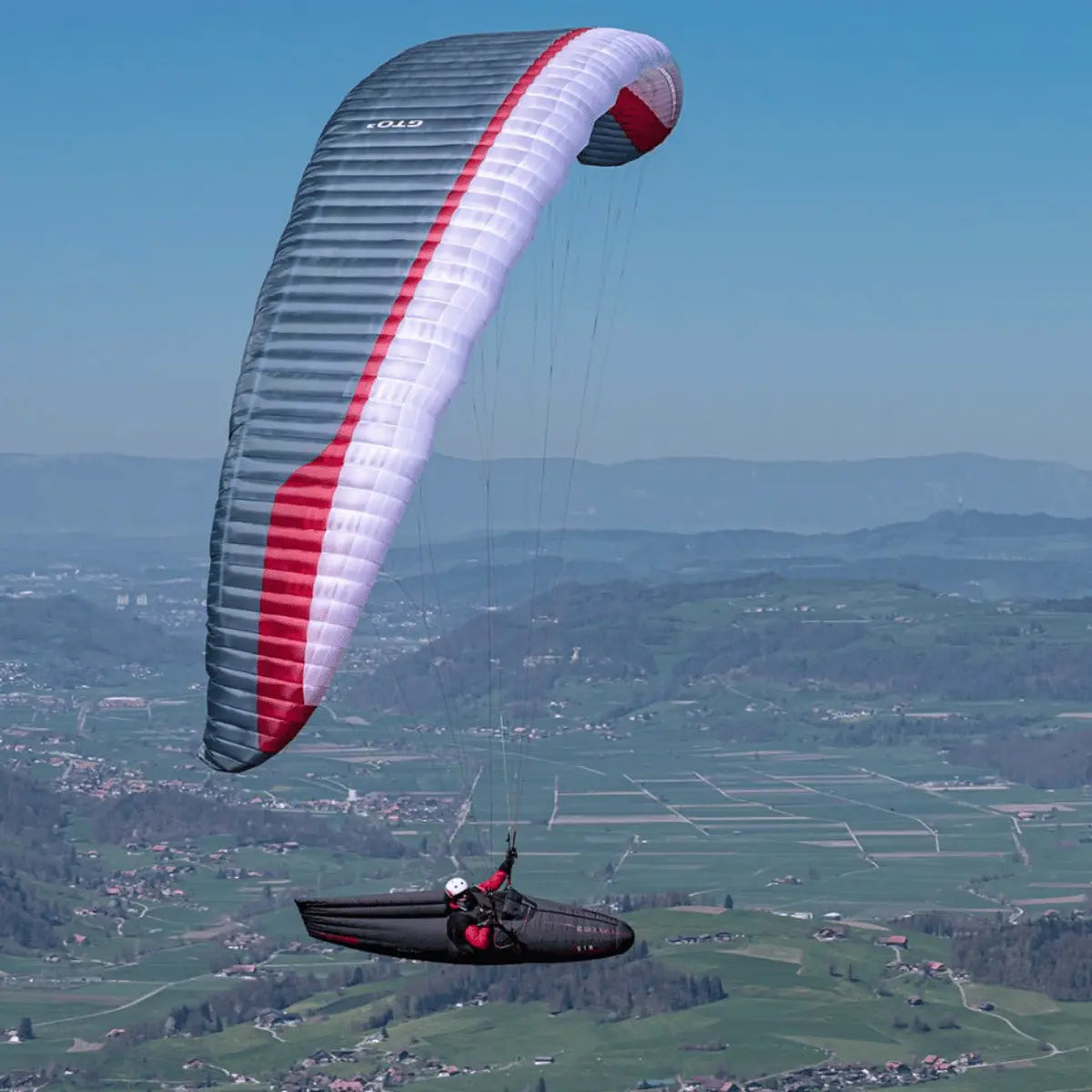 Person paragliding unter einem Gin GTO 3 EN-C, einer leistungsstarken EN-C Gleitschirmkappe, über grüner Landschaft. Ideal für erfahrene XC-Piloten.