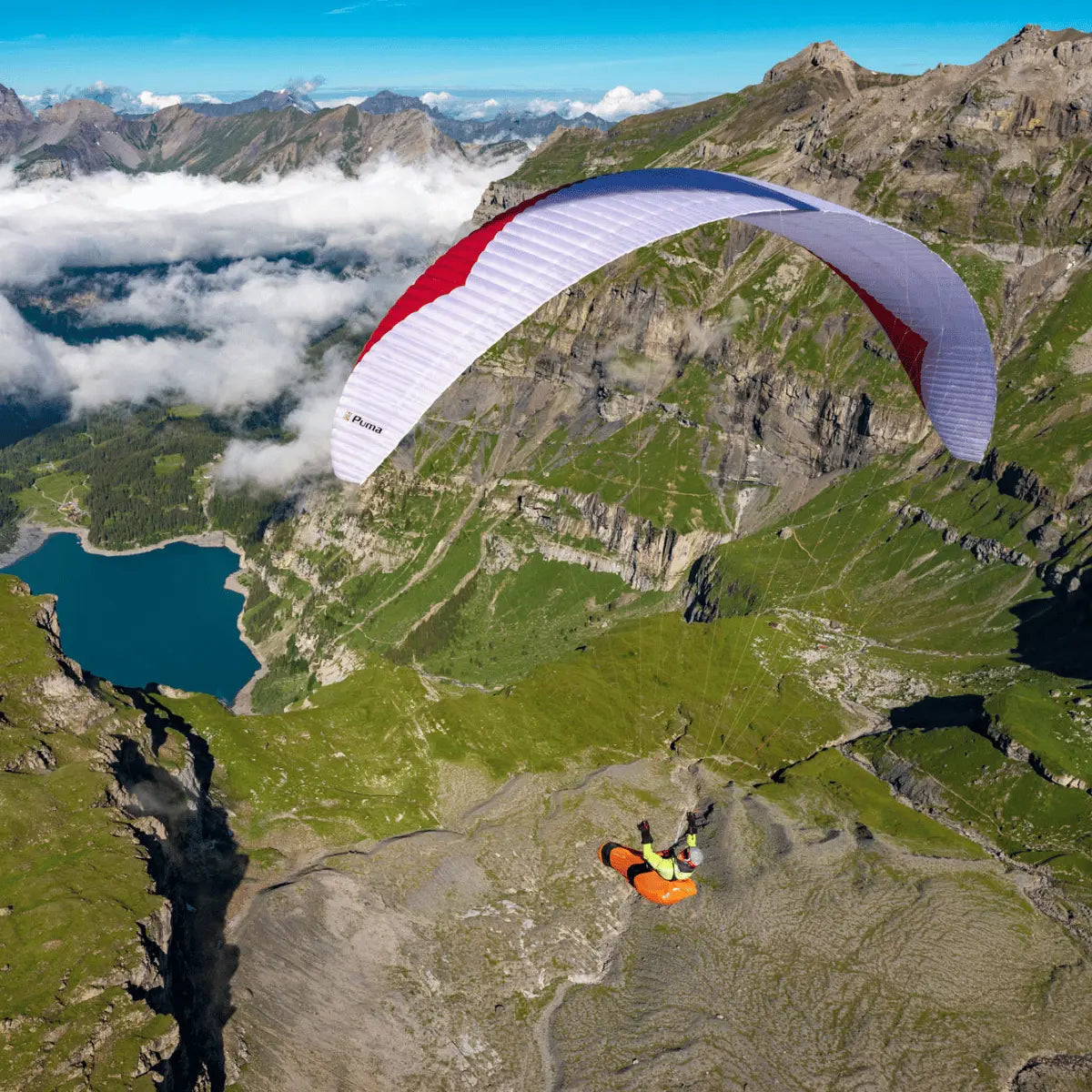 Paraglider Gin Puma Light (EN-D) über alpinem Gelände, Pilot in orangefarbenem Gurtzeug, weiße Flügel mit roten Akzenten, ideal für erfahrene XC- und Wettkampfpiloten.