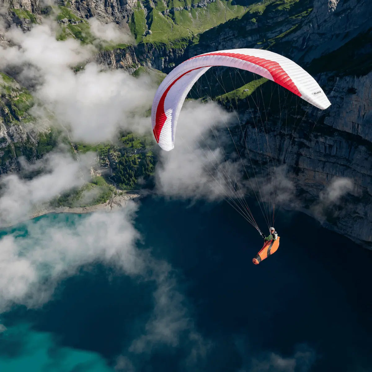 Person im Gurtzeug unter rotem Gin Puma Light (EN-D) Gleitschirm, schwebt über Bergsee und Klippen; ideal für erfahrene XC- und Wettkampfpiloten.
