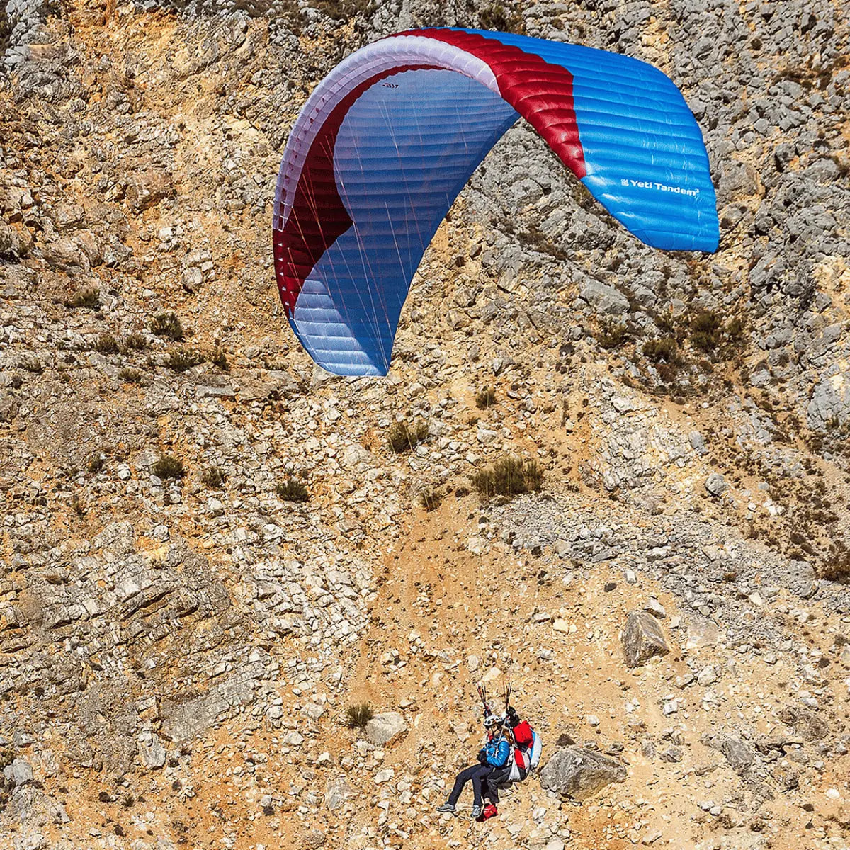 Zwei Personen fliegen mit dem Gin Yeti Tandem 3 (EN-A) Hike & Fly Tandem über eine felsige Landschaft, unter einem rot-weiß-blauen Schirm.