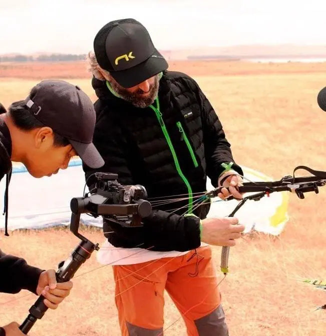 Niviuk Gleitschirm Down Jacket Pro, getragen von einem Mann im Freien, hält eine Kamera. Leichte und atmungsaktive Jacke für maximalen Flugkomfort.