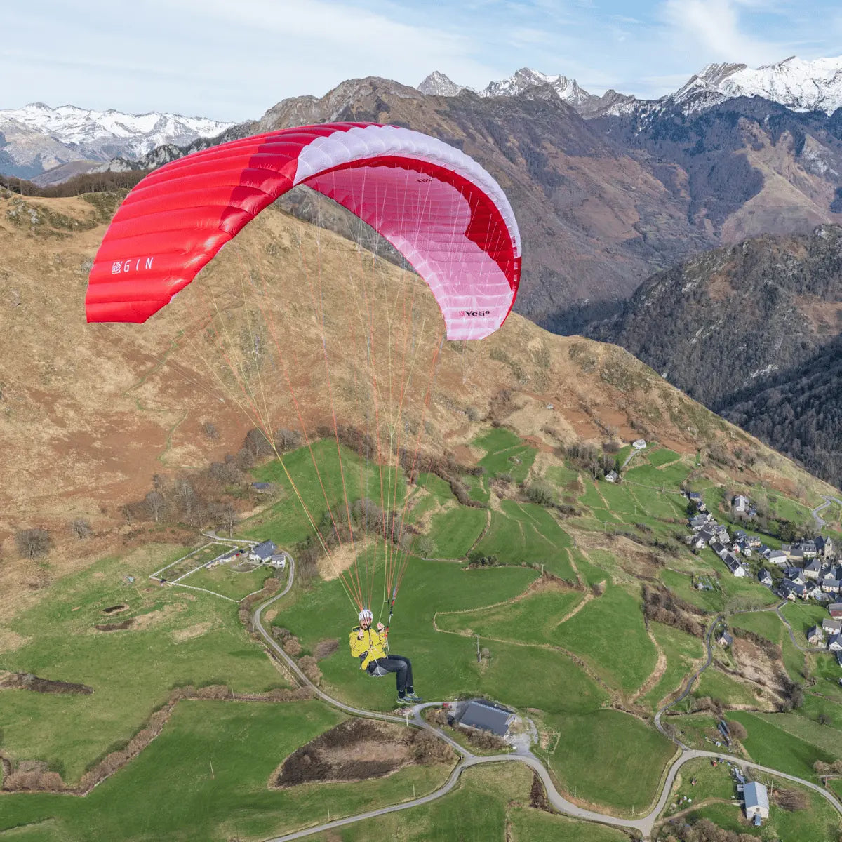Person fliegt mit Gin Yeti 6 Hike & Fly Gleitschirm über grüne Felder und Berglandschaft. Rotes, weiß-akzentuiertes Segel mit GIN Logo sichtbar.