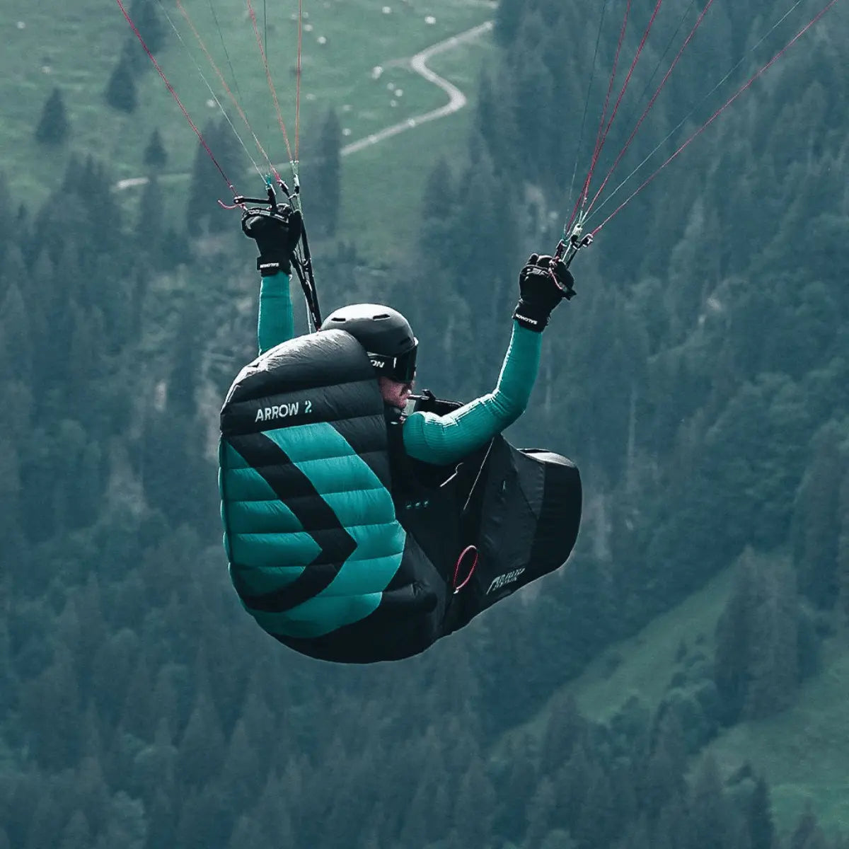 Person im Niviuk Arrow 2 Gurtzeug fliegt mit Gleitschirm über bewaldetem Berg, grün-schwarzes Design.