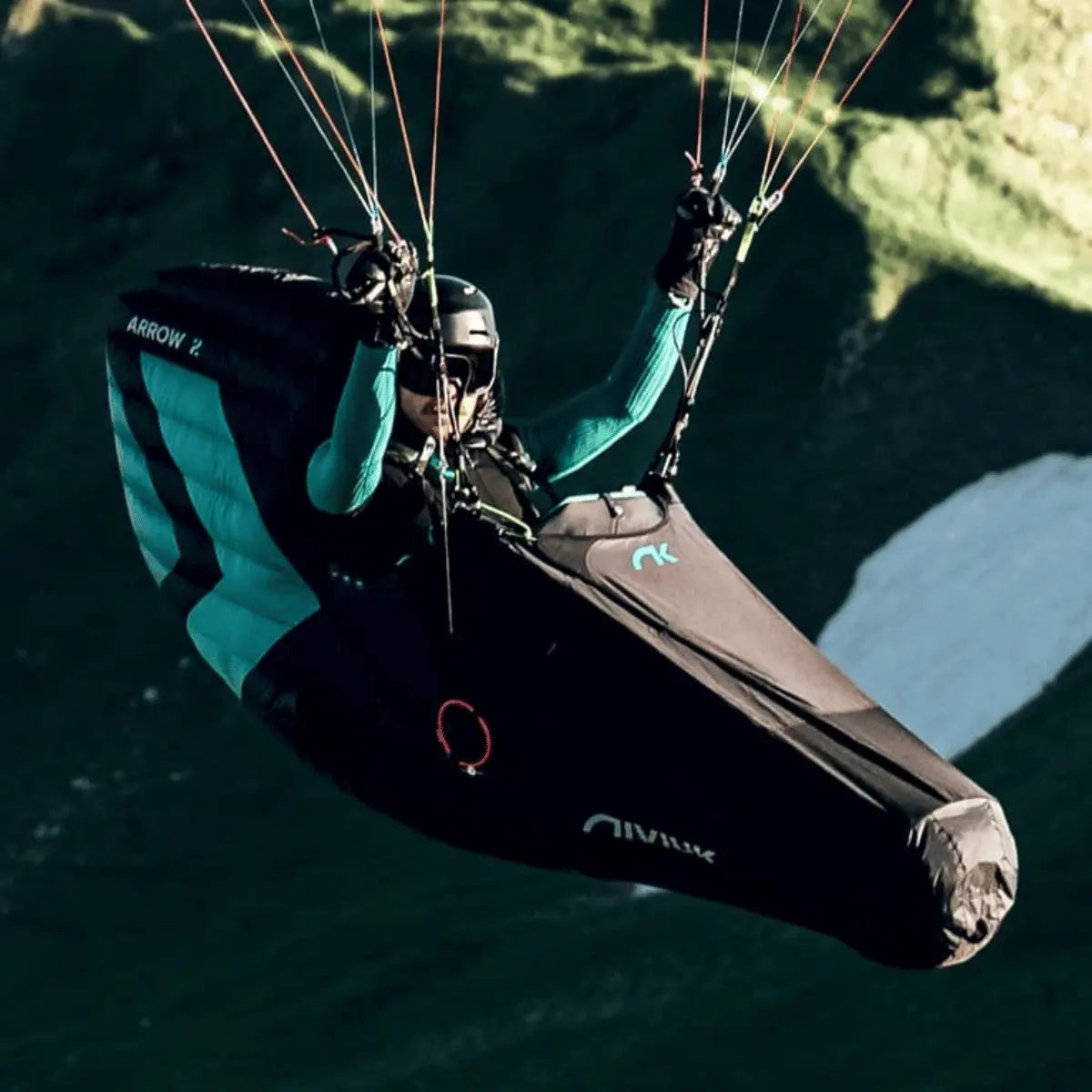 Niviuk Arrow 2 Liegegurtzeug für XC und Wettkampf in Schwarz und Türkis, geflogen von einer Person in der Luft über einer Berglandschaft.