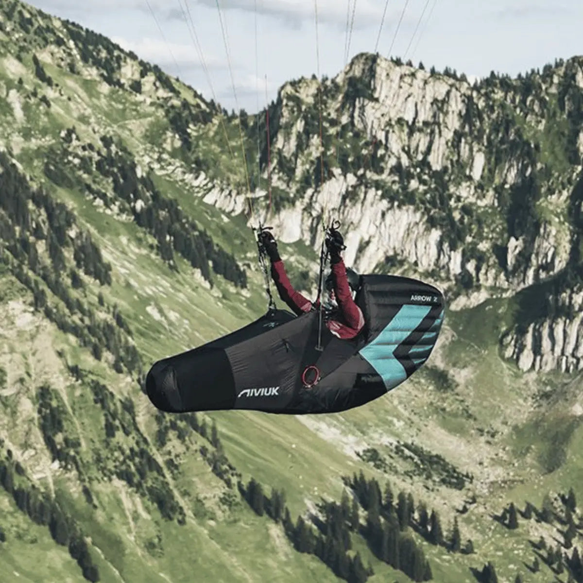 Niviuk Arrow 2 Liegegurtzeug in Schwarz mit türkisfarbenen Akzenten, beim Gleitschirmfliegen über grünen Bergen.