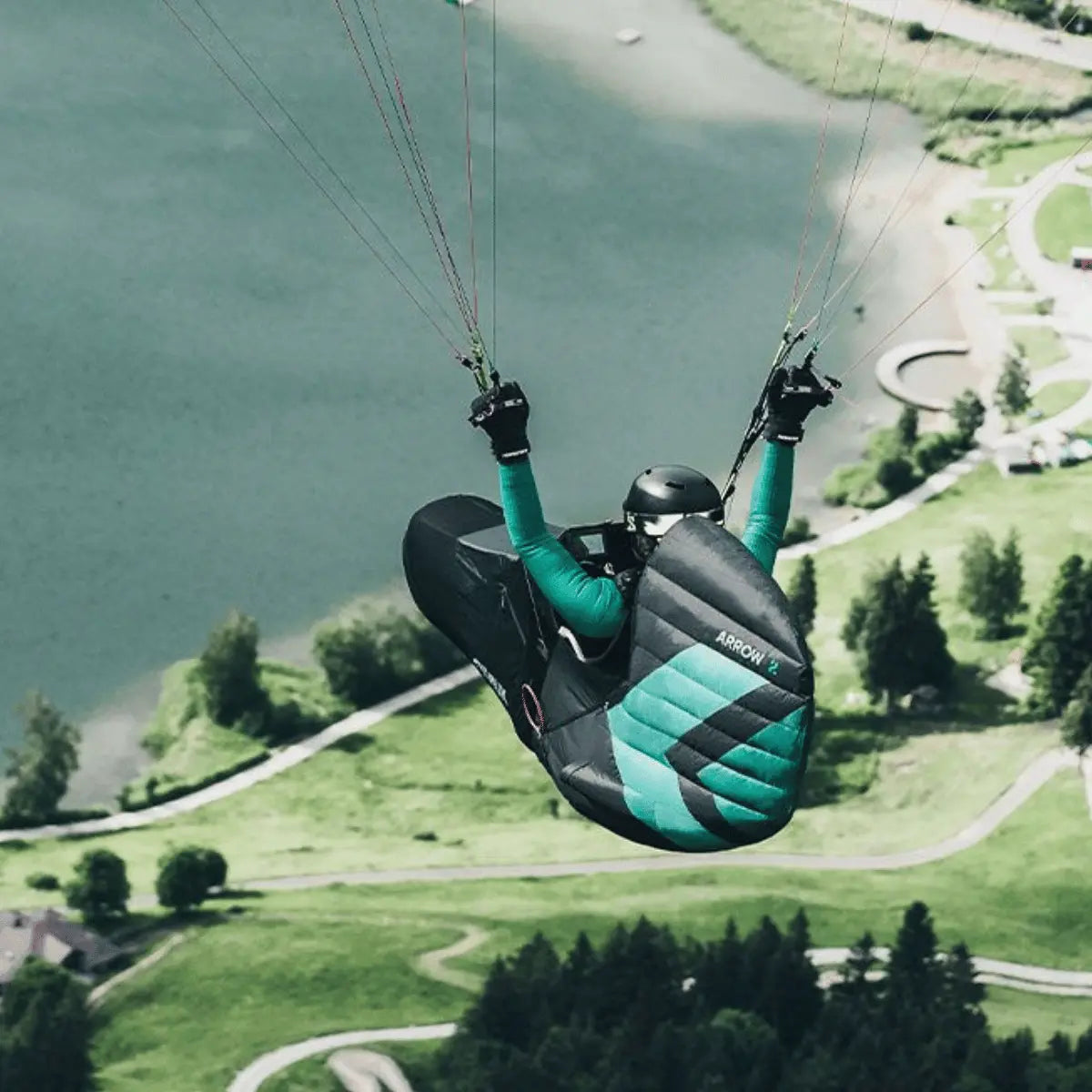 Niviuk Arrow 2 Gurtzeug im Flug, mit grünem Neopren-Einsatz und klar sichtbarem Logo, über einer grünen Landschaft mit See.