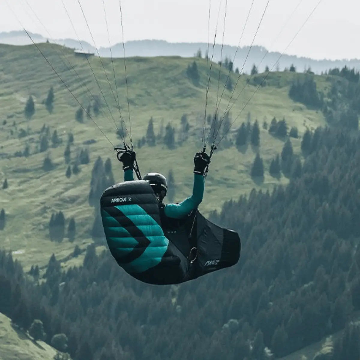 Ein Paraglider-Pilot in einem Niviuk Arrow 2 Liegegurtzeug fliegt vor einer grünen Berglandschaft mit Wäldern.