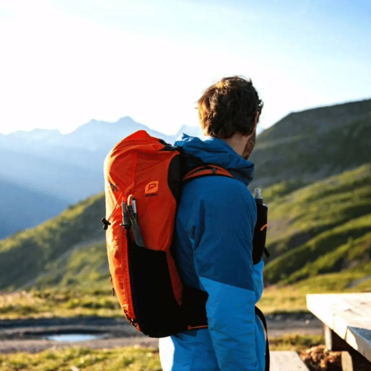 Orange Niviuk Expe Rucksack mit Flaschenhalter und Wanderstockhalter, getragen von einem Mann in blauer Jacke vor Bergkulisse.