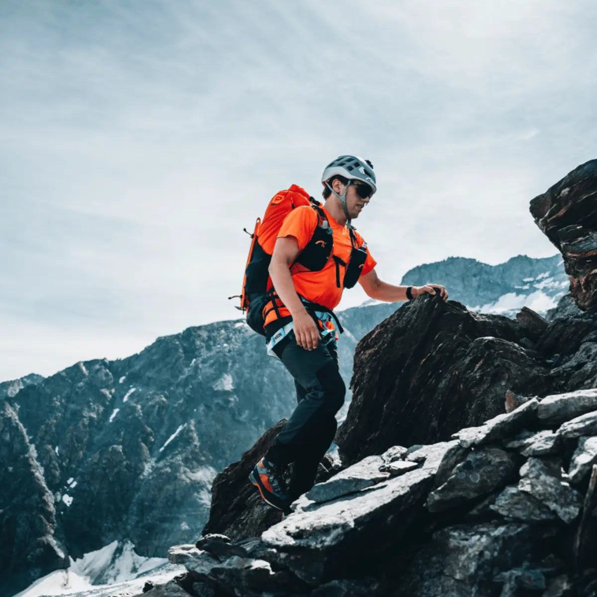 Ein Mann in orangefarbener Kleidung klettert mit einem roten Rucksack auf einem felsigen Berg.