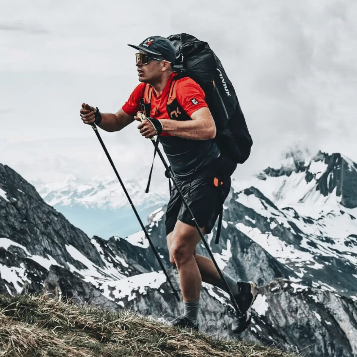 Ein Mann mit einem Niviuk Expe Race Rucksack wandert auf einem Bergpfad mit schneebedeckten Gipfeln im Hintergrund.