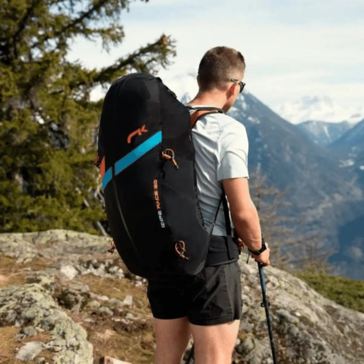 Ein Mann mit einem Niviuk Expe Race Rucksack auf dem Rücken steht in den Bergen und blickt in die Ferne. Der schwarze Rucksack hat orangefarbene und blaue Akzente.
