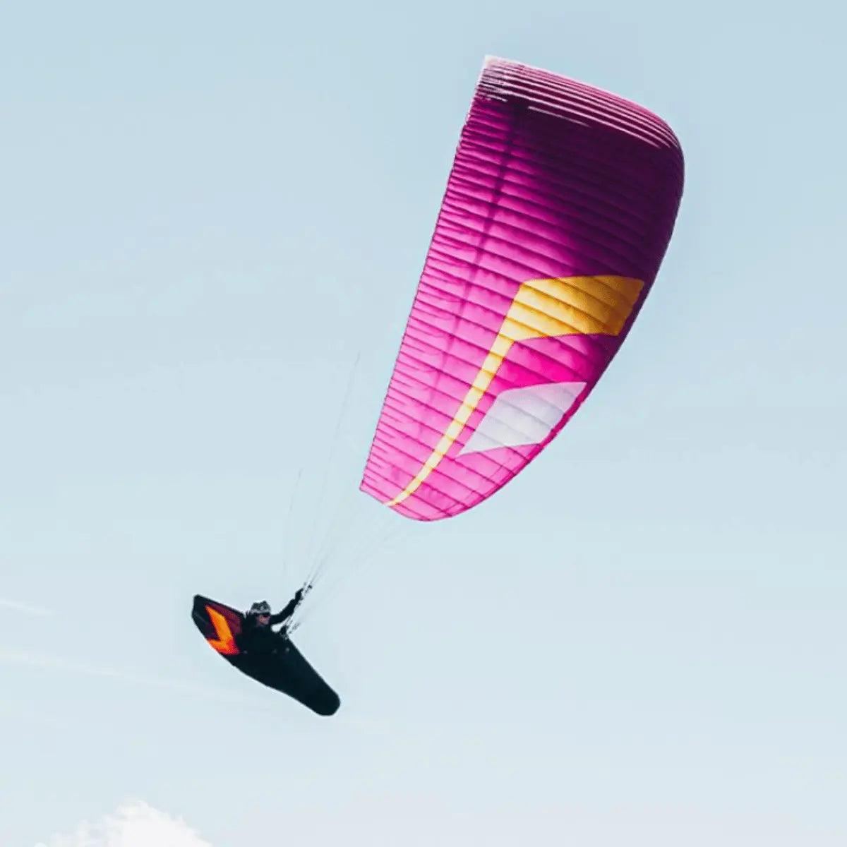 Ein pinker Niviuk Hiko P Gleitschirm mit gelben und weißen Akzenten fliegt mit einem Piloten im Trapez gegen einen blauen Himmel.