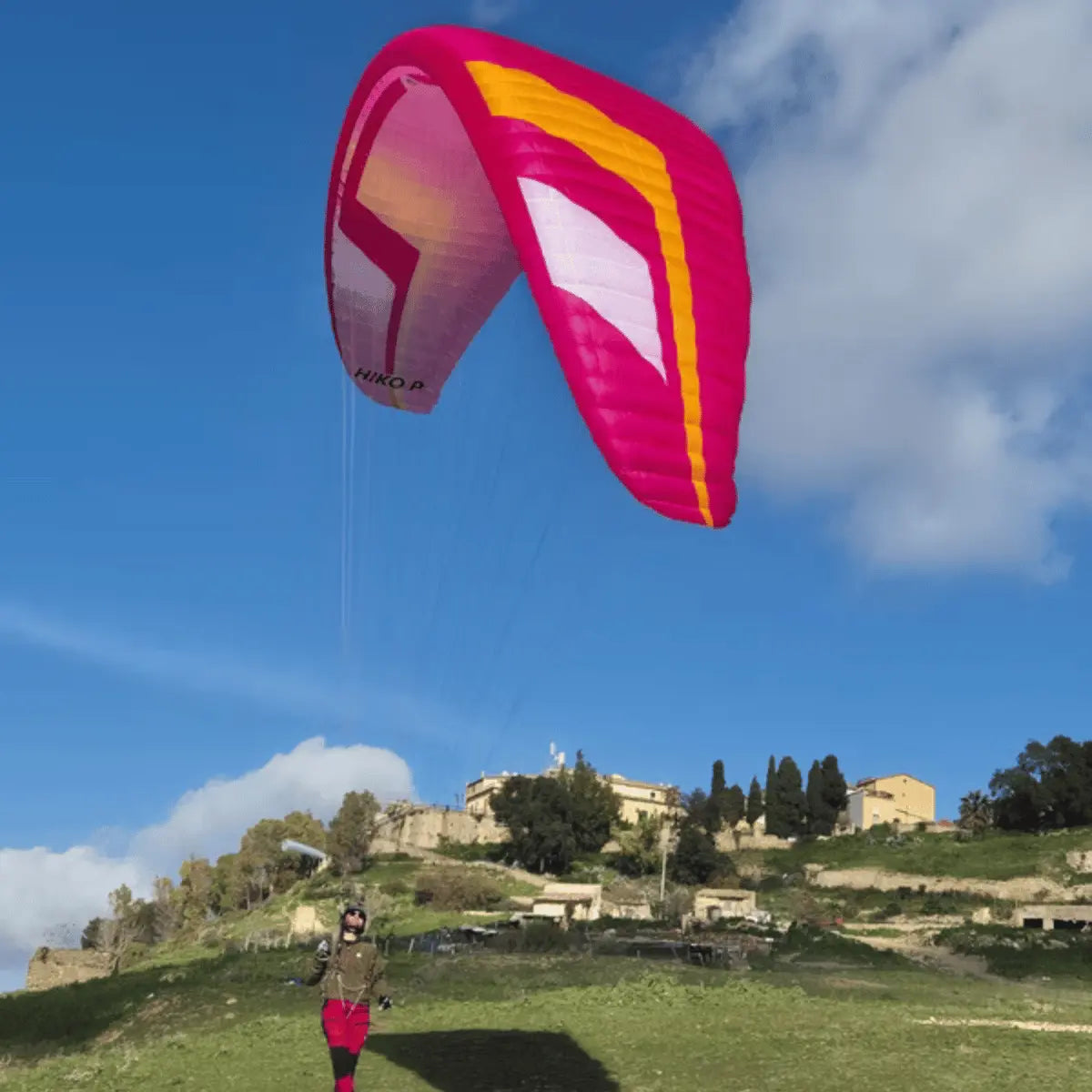 Ein pink-gelber Niviuk Hiko P Gleitschirm fliegt über einer grünen Hügellandschaft mit Gebäuden im Hintergrund.