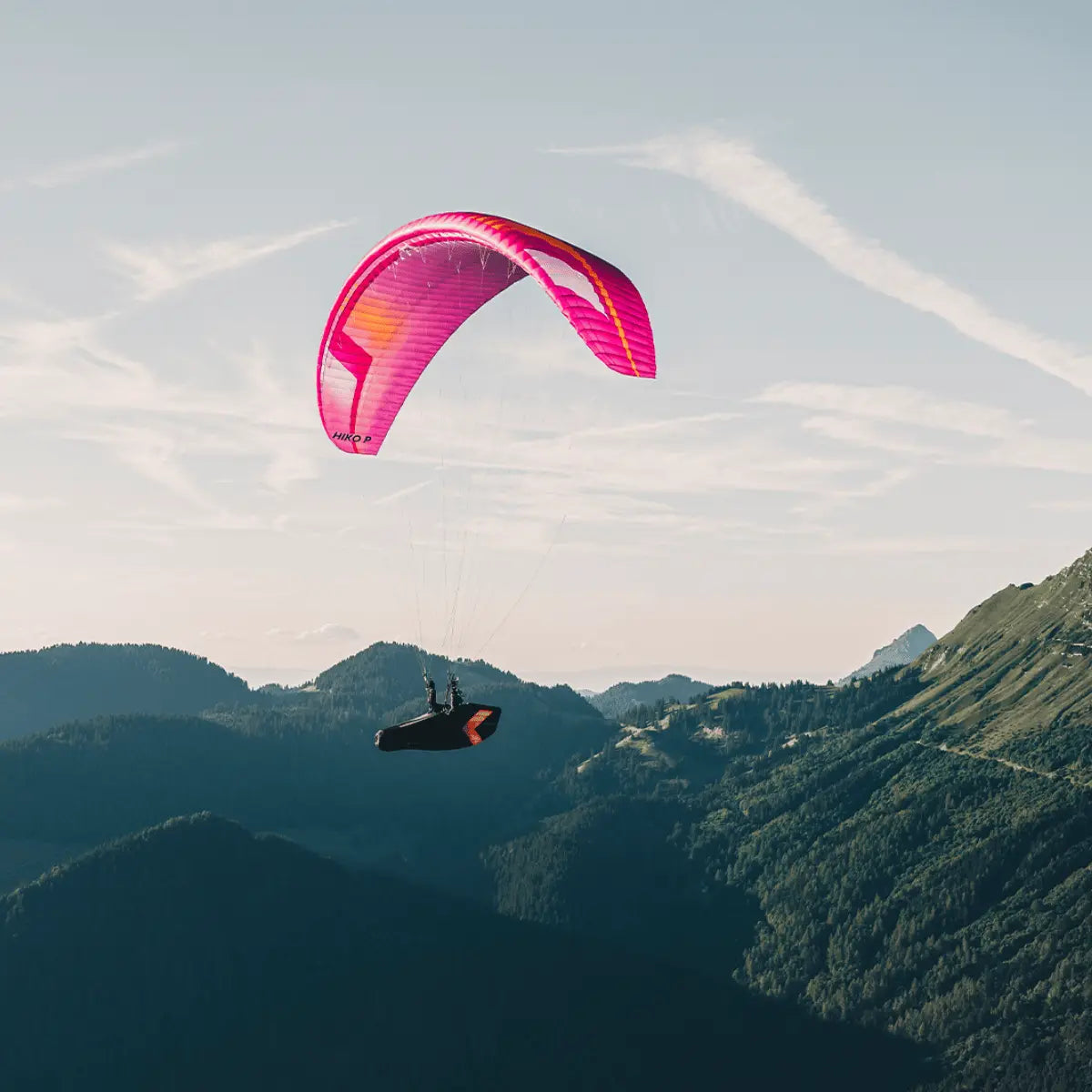 Pink Niviuk Hiko P Gleitschirm mit orangen Akzenten, der von einem Gleitschirmpiloten über einer Berglandschaft geflogen wird.