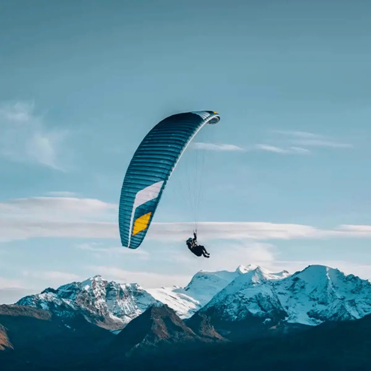 Ein Gleitschirmflieger genießt den Flug unter einem blauen und gelben Niviuk Hiko P Gleitschirm mit schneebedeckten Bergen im Hintergrund.