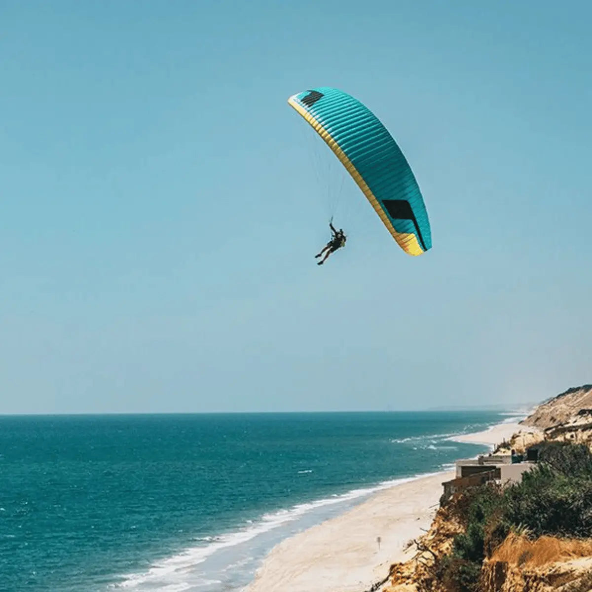 Ein Mann fliegt mit einem blau-gelben Niviuk Jester Paragleiter über einer Küstenlandschaft mit dem Meer und Strand.