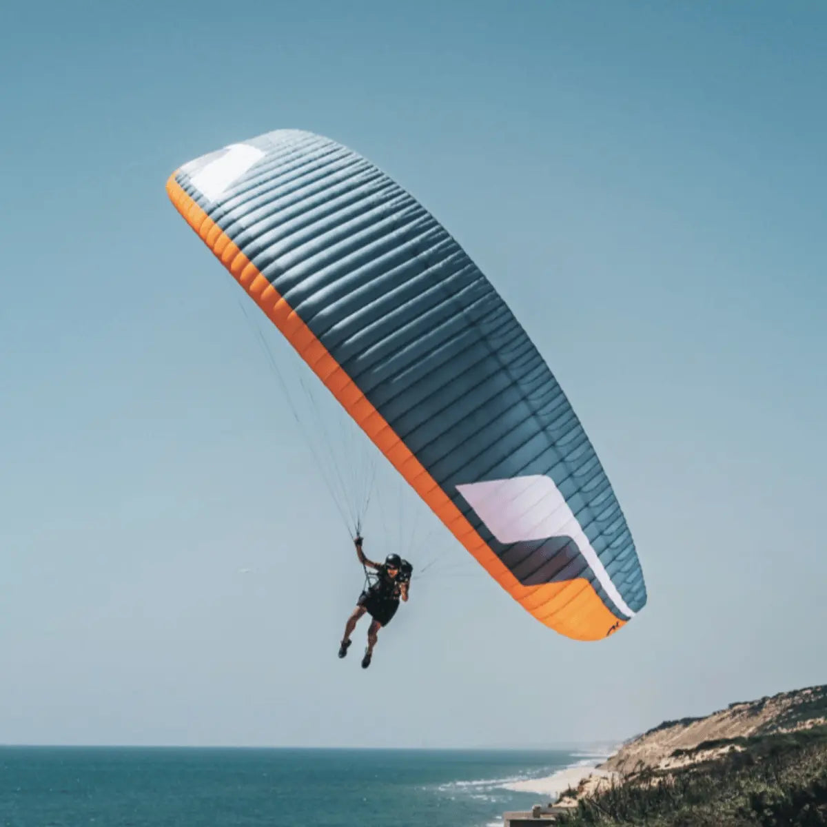 Ein Paragleiter im Flug, mit einem Piloten, der unter dem orange-blauen Schirm hängt, vor der Küste und dem Meer.