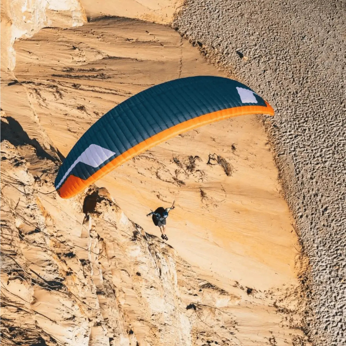 Ein Niviuk Jester Gleitschirm mit orangefarbenem Rand und blauem Oberteil fliegt über sandige Dünen, gesteuert von einem Paragleiter.