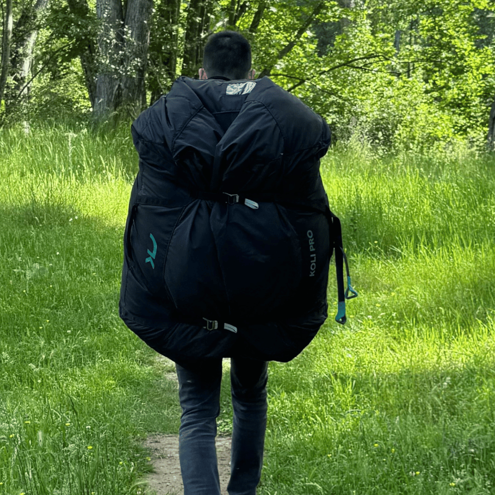 Niviuk Koli Pro Tandem Schnellpacksack, getragen von einem Tandempiloten auf einem Naturweg im Wald. Ideal für schnelles, sicheres Packen von Flugausrüstung, mit elastischem Kordelzug und verstellbaren Trageriemen.