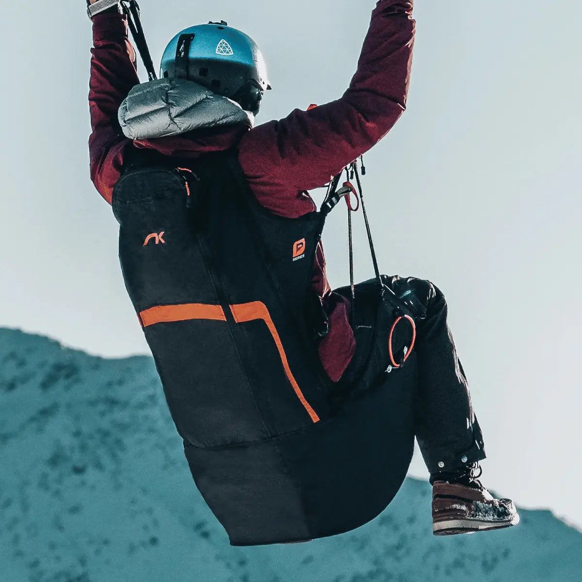 Ultraleichtes Wendegurtzeug Niviuk Kooper P mit orangefarbenen Akzenten, getragen von einem Gleitschirmpiloten mit Helm, der im Flug schwebt.