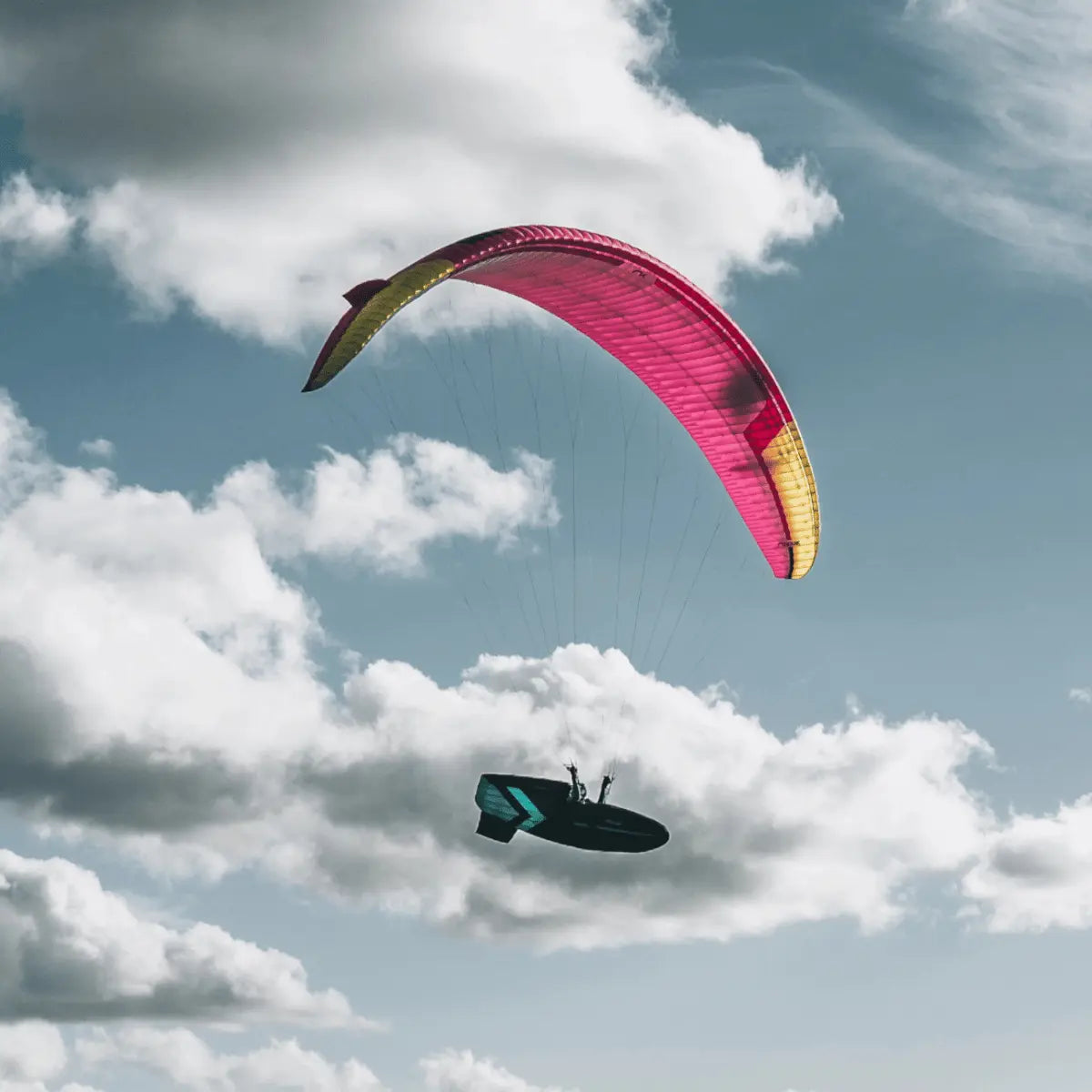 Ein Niviuk Peak 7 Hochleistungs-Gleitschirm in Pink und Gelb fliegt hoch am Himmel mit einem Piloten im Gurtzeug unter einer schwarzen Kappe.