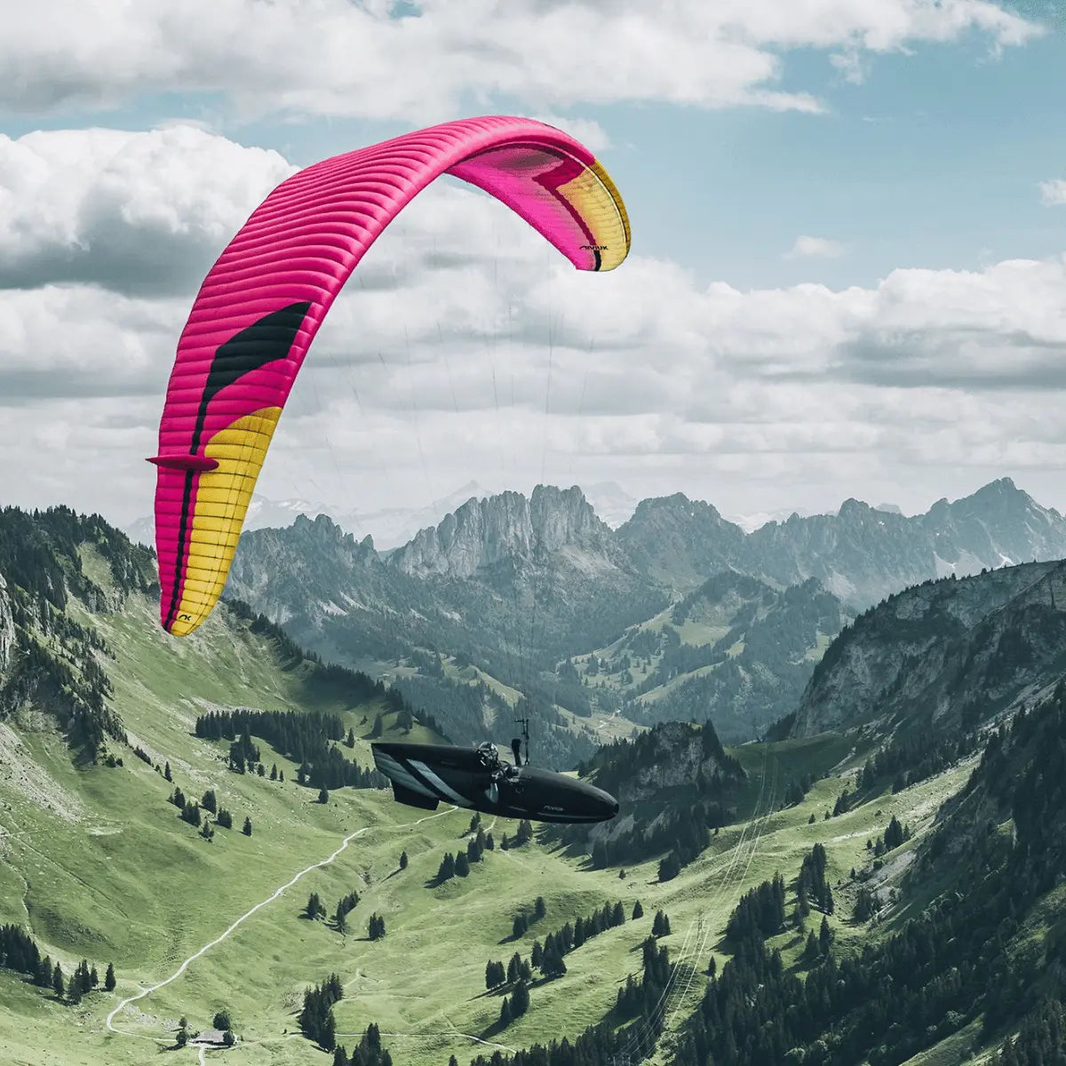 Niviuk Peak 7 (EN-D) Hochleistungs-Gleitschirm in Pink und Gelb fliegt über einer alpinen Berglandschaft mit einem Piloten im Gurtzeug.