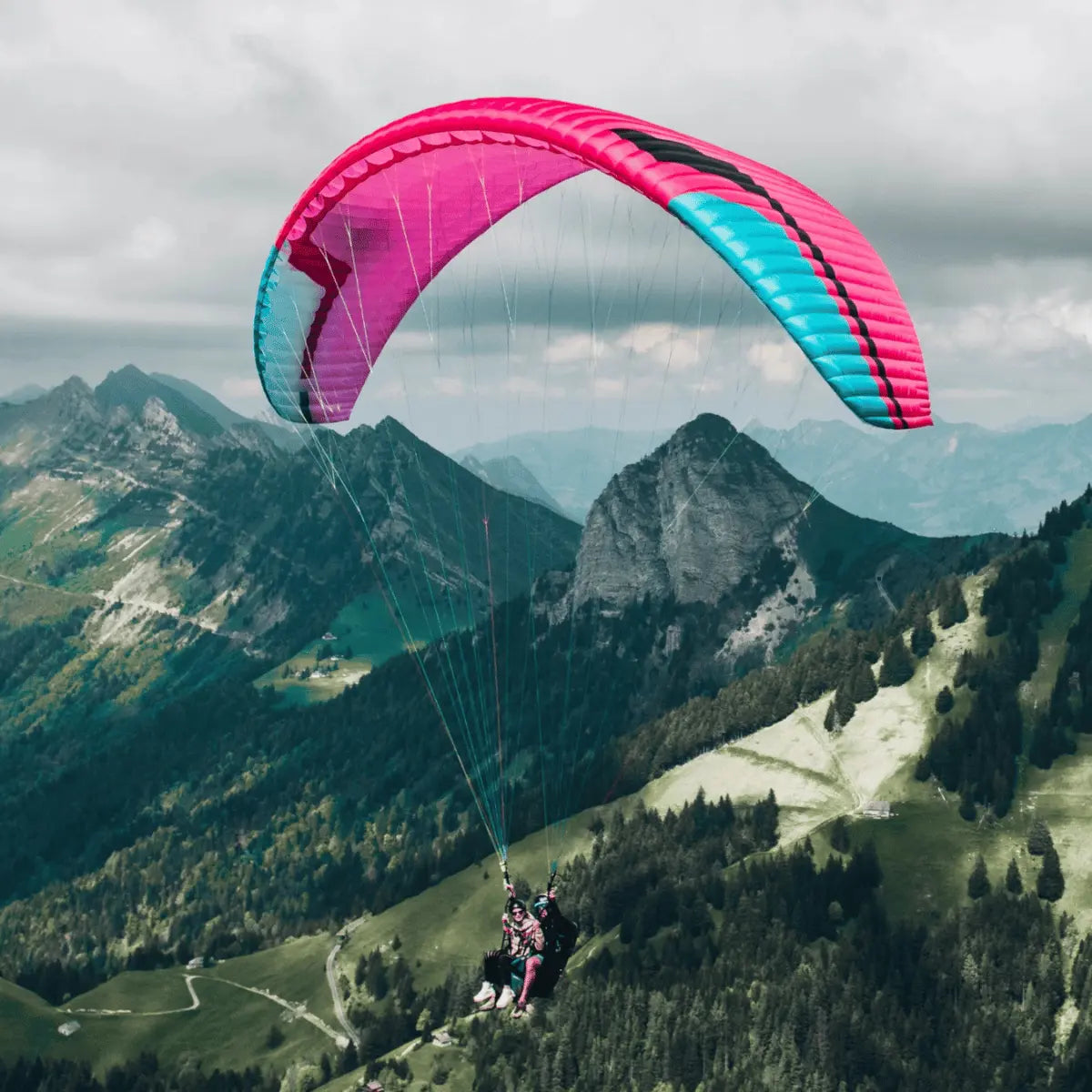 Zwei Personen schweben mit einem pink-blauen Tandem-Gleitschirm von Niviuk über einer Berglandschaft.