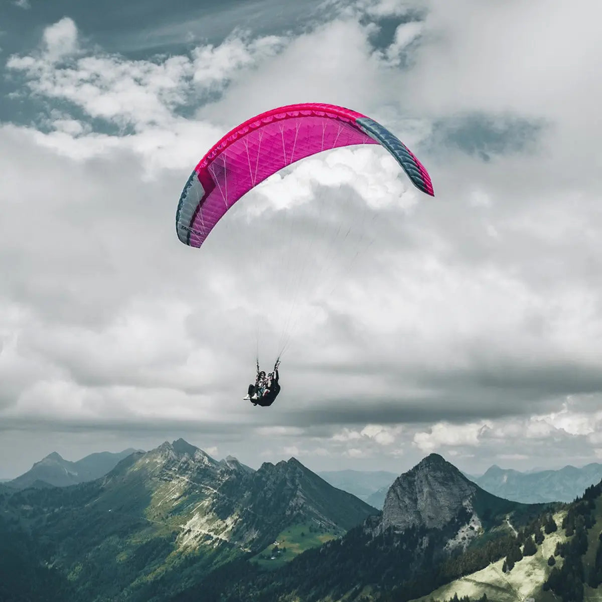 Zwei Personen fliegen mit einem pink-grauen Tandem-Gleitschirm Niviuk Takoo 6 über einer Berglandschaft.