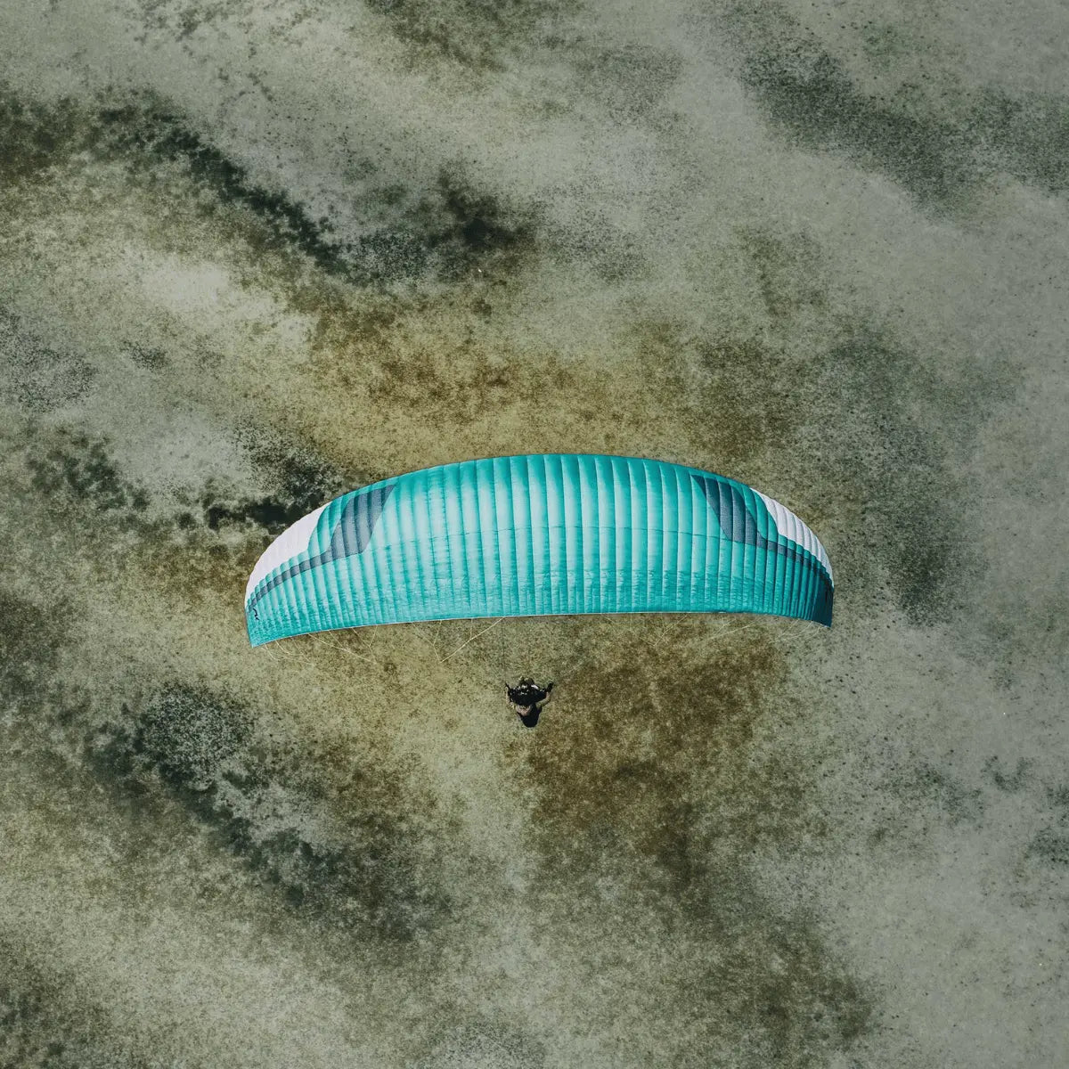 Türkisfarbener Tandem-Gleitschirm Niviuk Takoo 6 (EN-B) mit weißem Streifen fliegt über eine erdige Landschaft.