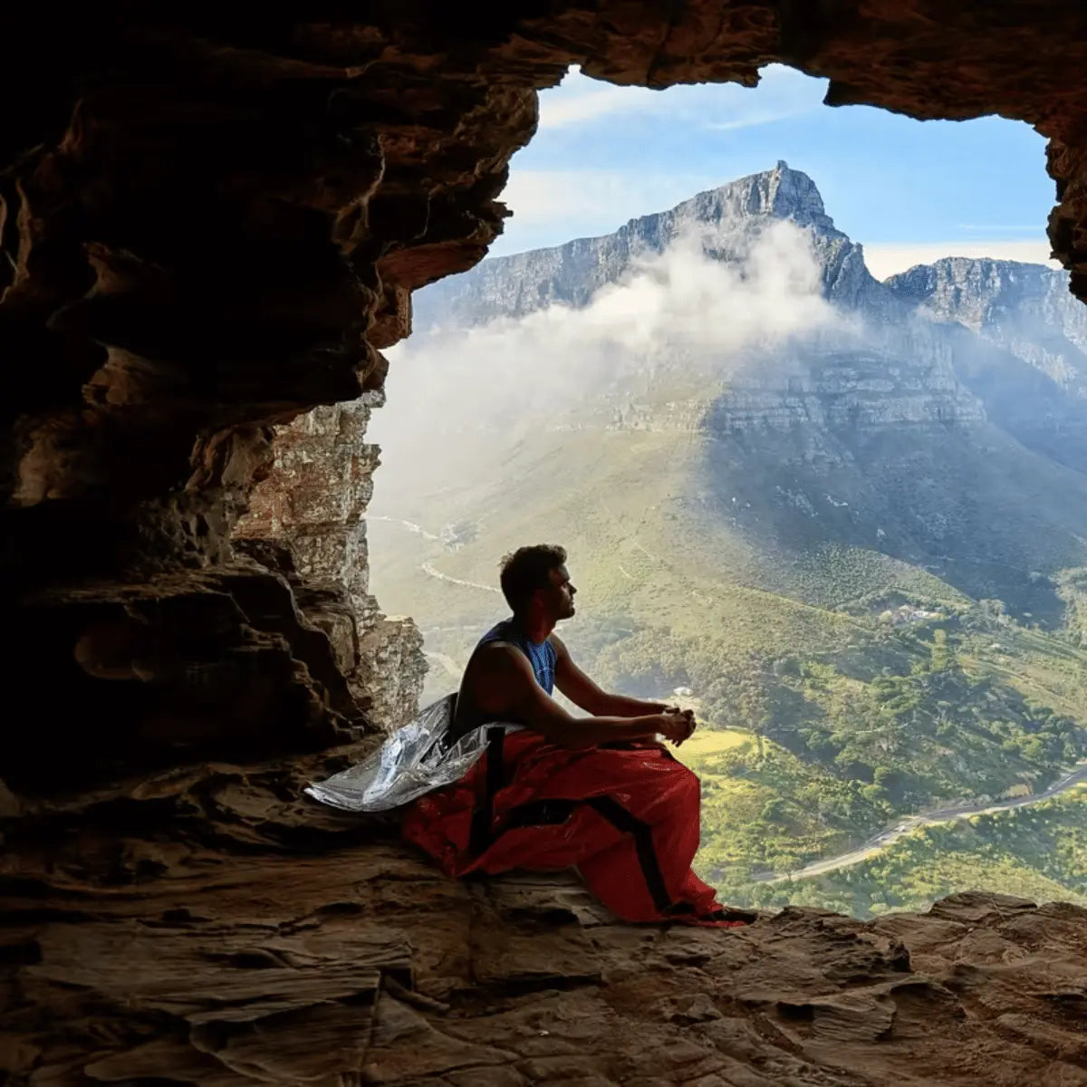 Mann in Notfall Bivy Schlafsack sitzt in Höhle mit Blick auf Berglandschaft.