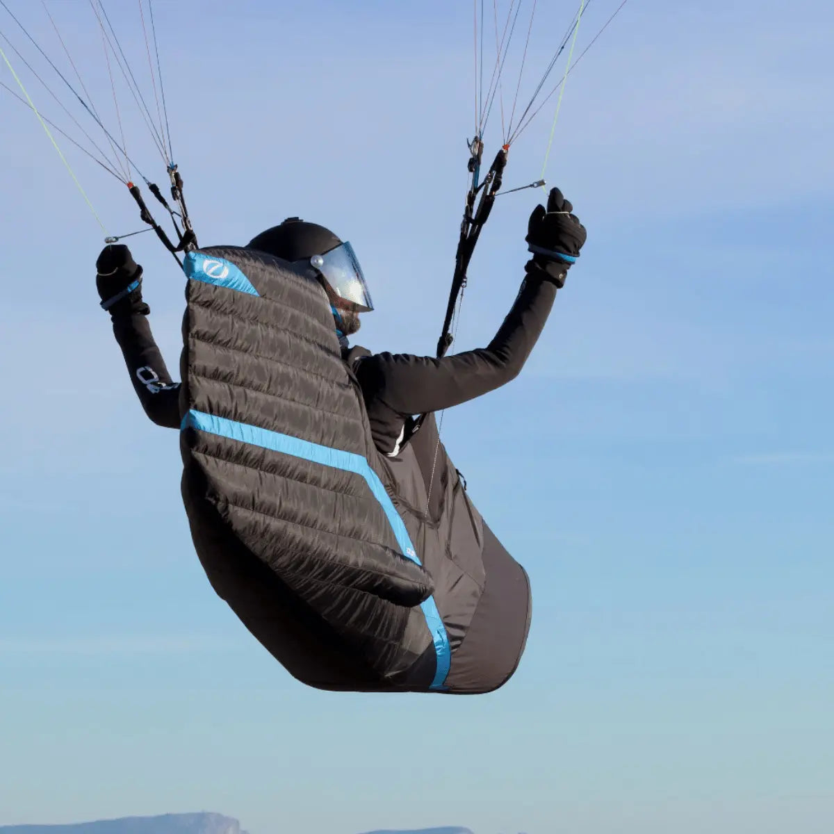 Der Ozone Ozium 3 Pod Liegegurtzeug in Schwarz mit blauen Akzenten wird von einem Paraglider in der Luft getragen.
