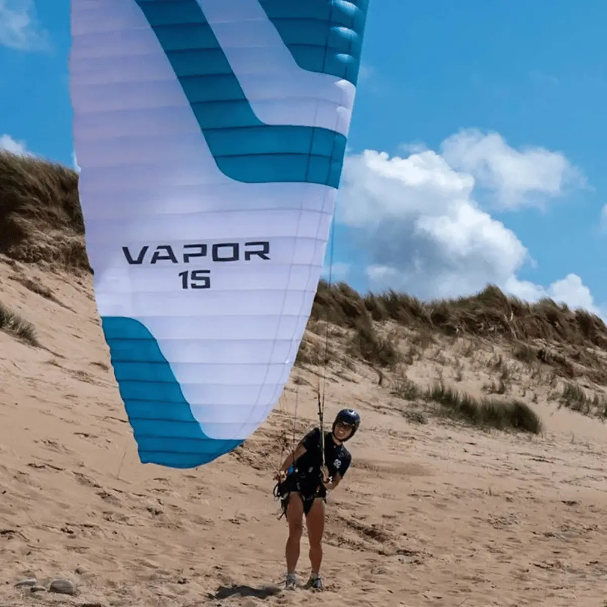 Ein weiß-blauer Ozone Vapor 15 Parakite mit Streifen, der auf sandigem Untergrund mit einer Person im Geschirr steht, vor einem bewölkten Himmel.