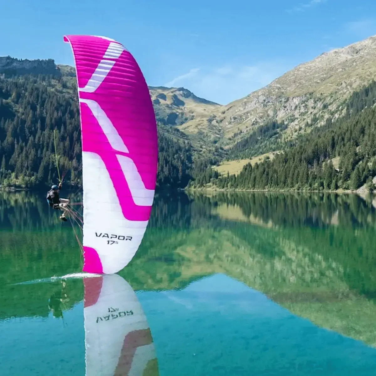 Ein pink-weißes Gleitschirm-Segel mit dem Schriftzug "VAPOR 17" schwebt über einem spiegelglatten Bergsee, ein Gleitschirmpilot ist angeschnallt.