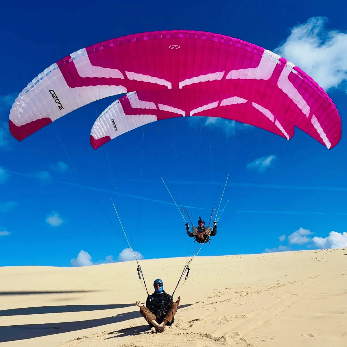 Zwei Parakites, ein großer pink-weißer Ozone Vapor und ein kleinerer, fliegen über einer Sanddüne unter blauem Himmel.