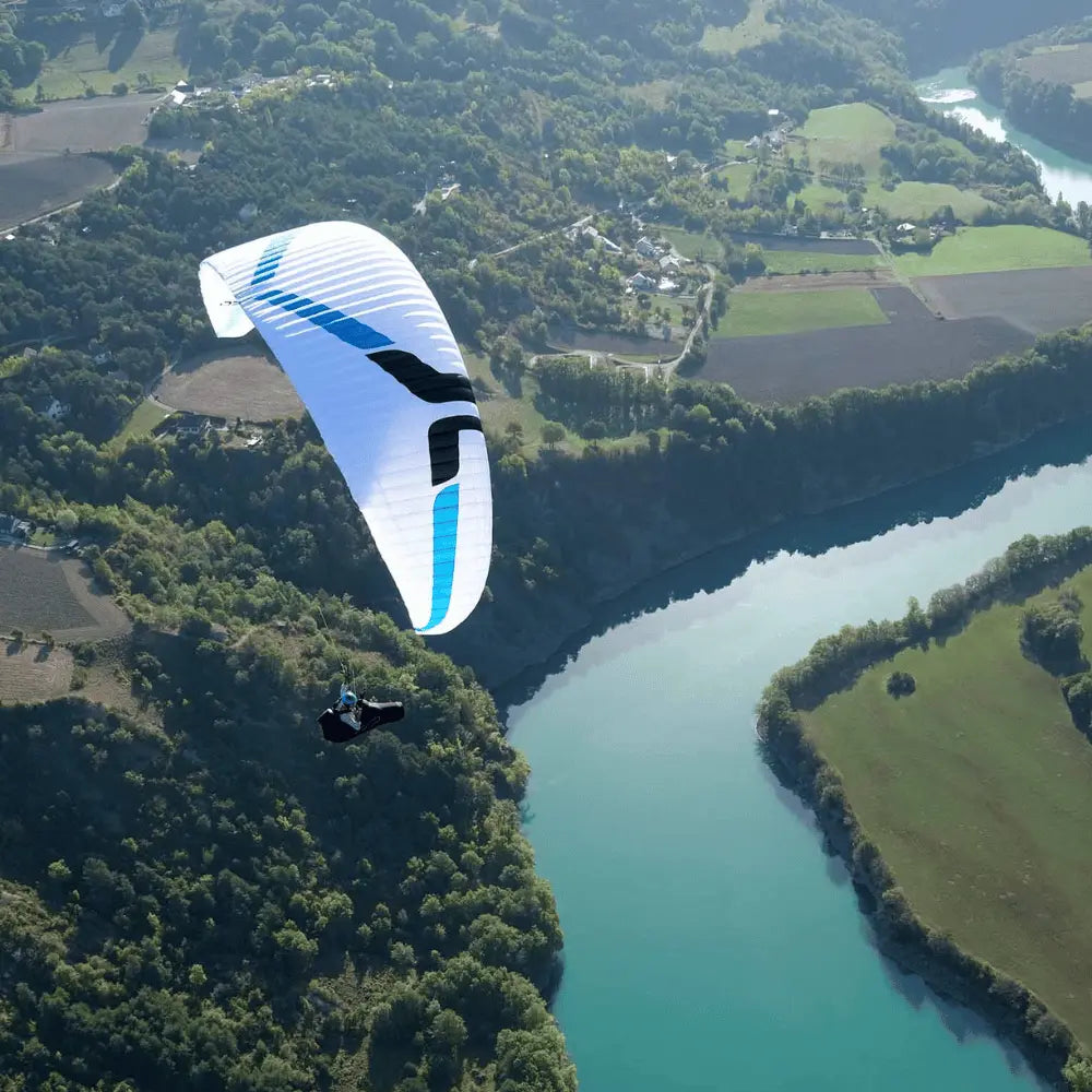 Person beim Paragliding mit weissem Ozone Rush 6 über grüner Landschaft, symbolisiert hohe Leistung und Stabilität im Flug, passend für erfahrene Freizeit- und XC-Piloten.