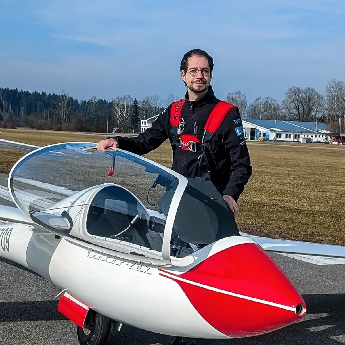 Mann in Segelflieger-Kleidung steht neben einem roten und weißen Segelflugzeug auf einem Flugplatz.