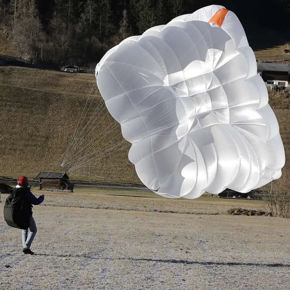 Person hält Charly Targetcross ST Light Notschirm mit weißem Schirm und orangefarbener Spitze, ideal für sicherheitsbewusste Gleitschirmflieger.