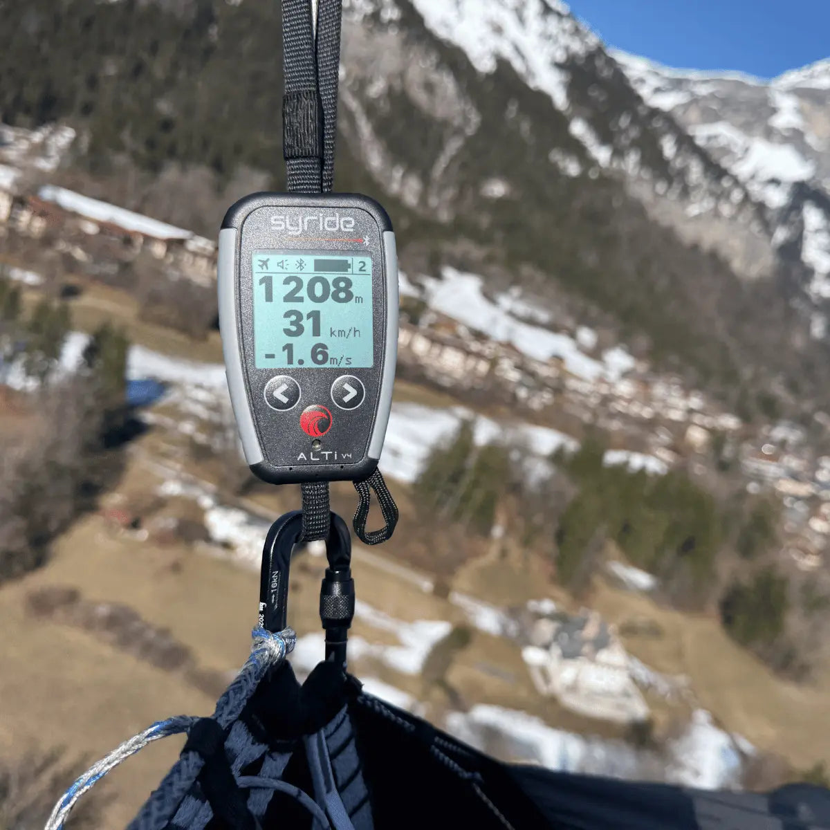 Syride Alti V4 mit digitalem Display zeigt Höhe, Geschwindigkeit und Steigrate an, befestigt an einem Karabiner über verschneiter Berglandschaft